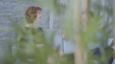 Oldukça zarif olgun kadın yakın kanepede oturan mavi gömlekli genç adam ile konuşurken. Bitki yaprakları ön planda görünümü kapsamaktadır. Yaşlı kadın ve genç erkek arasındaki ilişki