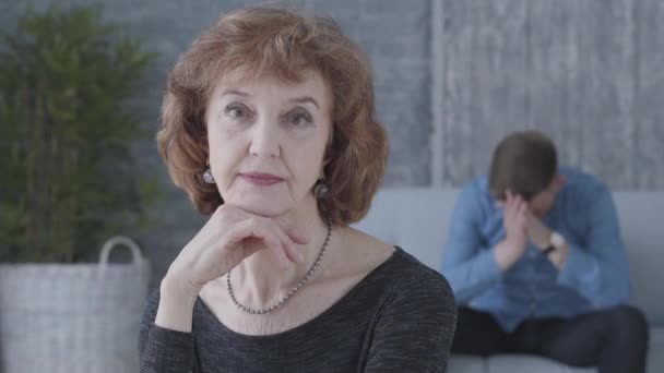 Portrait of a senior mature woman looking in the camera in the foreground. The blurred figure of the sad unhappy man sitting on the sofa on the background. The old lady has a quarrel with her young — Stock Video