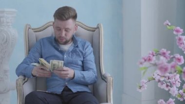 Mavi gömlekli mutlu zengin sakallı adam ışık odasında beyaz koltukta oturup para sayıyor. Elinde dolar olan genç bir işadamı. Kamera sola hareket ediyor