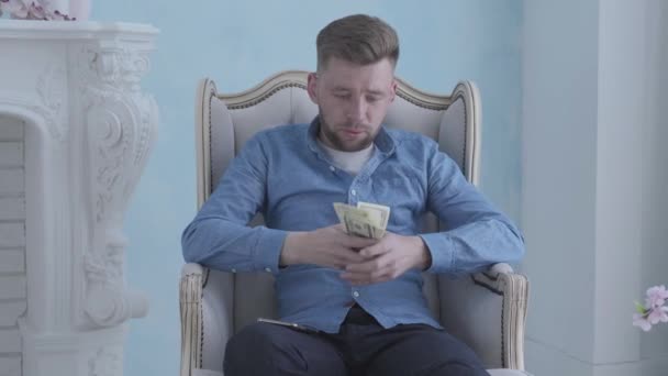 Feliz hombre barbudo rico reflexivo en camisa azul sentado en el sillón blanco en la sala de luz contando dinero. Joven hombre de negocios con dólares en las manos. Cámara moviéndose derecha — Vídeo de stock