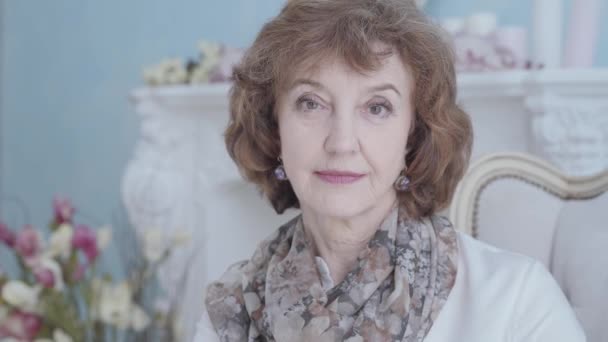 Happy positive elegant mature lady sitting in the armchair looking in the camera close up. Flowers in the background. Camera moving right — Stock Video