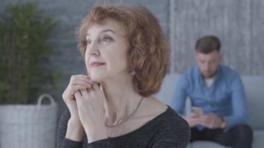 Ön planda üst düzey bir olgun kadının portresi kadar kapatın. Arka planda kanepede oturan üzgün mutsuz adamın bulanık figürü. Yaşlı kadın genç kocasıyla tartışıyor.