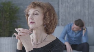 Ön planda kameraya bakan üst düzey bir olgun kadının yakın portresi. Arka planda kanepede oturan üzgün mutsuz adamın bulanık figürü. Yaşlı kadın onunla tartışıyor.