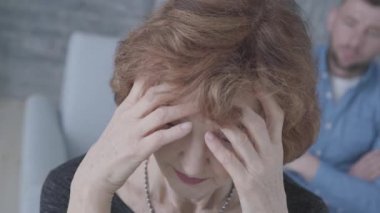 Üst düzey olgun zarif üzgün kadın ön planda başını tutarak yakın yüzü. Arka planda kanepede oturan sinirli adamın bulanık figürü. Yaşlı kadın genç kocasıyla tartışıyor.