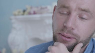 Yakın portre yakışıklı olumlu gülümseyen sakallı adam mavi gömlek beyaz mobilya ile odada koltukta oturan. Kamera sağa hareket ediyor
