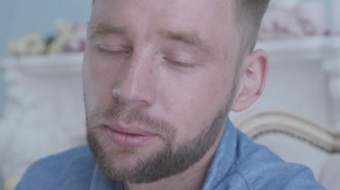 Yakın yüz yakışıklı pozitif gülümseyen sakallı adam mavi gömlek beyaz mobilya ile odada koltukta oturan. Kamera sağa hareket ediyor