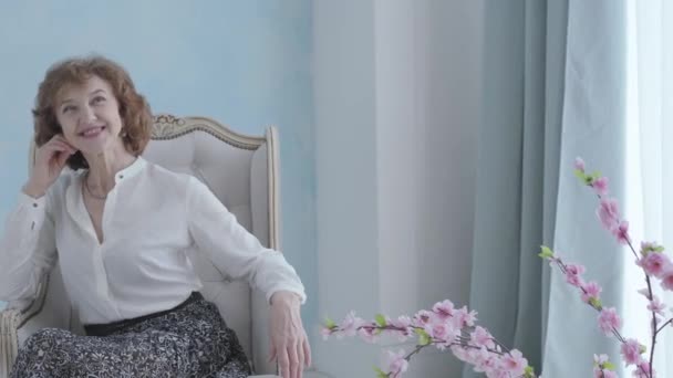 Portrait belle femme mature élégante et mignonne en chemisier blanc assis dans le fauteuil souriant. L'homme méconnaissable debout le dos à la caméra tenant un bouquet de roses roses pour sa dame — Video