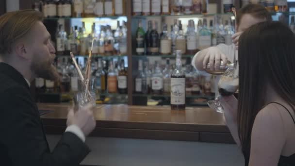 Mladý pár sedí v baru v drahé restauraci nebo hospodě. Vousatý sebevědomý muž pije whisky a jeho přítelkyně pije koktejl. Barman připraví drink v baru.. — Stock video