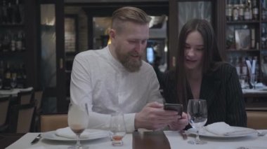 Sarışın sakallı adam ve masadaki restoranda oturan güzel esmer kadın. Adam kız arkadaşına cep telefonundan bilgi gösteriyor. Sevimli çift bir tarih var