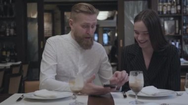 Yakışıklı sakallı adam ve güzel esmer kadın masada restoranda oturuyor. Adam kız arkadaşına cep telefonundan bilgi gösteriyor. Sevimli çift bir tarih var