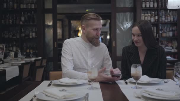 Attrayant homme barbu et jolie femme brune assise dans le restaurant à la table. L'homme montre à sa copine des informations sur le portable. Couple mignon a une date — Video