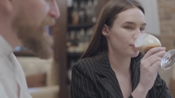 Glamour brunette vrouw drinken cocktail zittend aan de tafel met haar bebaarde vriendje luisteren naar hem. Het schattige paar heeft een date. Camera links en rechts bewegen — Stockvideo