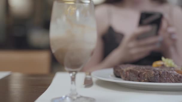 Вкусный стейк с овощами и листьями лежит на тарелке перед женщиной в ресторане. Лэдис пишет смс-ки на мобильный. Алкогольное стекло стоит рядом. Камера движется вправо — стоковое видео