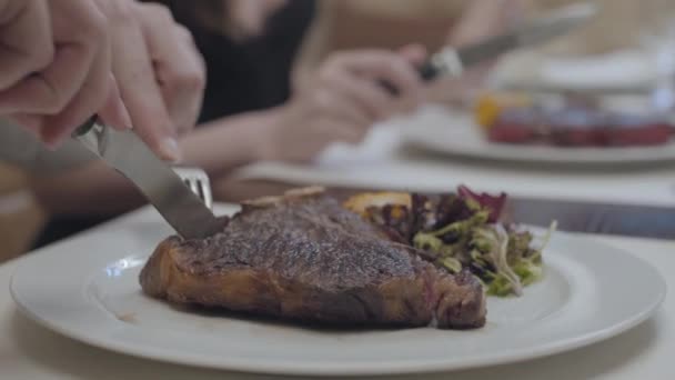 Close up hands of young man in the foreground and woman in the background cutting tasty steak lying on the plate with leaves and vegetables in the restaurant. The cute couple has a date. — Stock Video