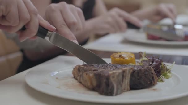 Die Hände des jungen Mannes im Vordergrund und der Frau im Hintergrund schneiden leckere Steaks, die auf dem Teller liegen, mit Blättern und Gemüse in der Nähe des Restaurants. Das süße Paar hat ein Date. — Stockvideo