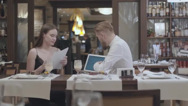 Portret jonge mooie vrouw zitten met papieren op de tafel in restaurant met een zelfverzekerde bebaarde man die haar informatie over de laptop te tonen. Zakelijk diner. — Stockvideo