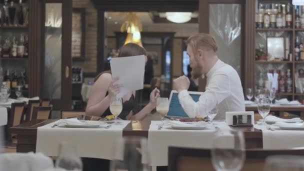 Portret schattige jonge vrouw zitten met papieren op de tafel in restaurant met een zelfverzekerde bebaarde man die haar informatie over de laptop te tonen. Zakelijk diner. — Stockvideo