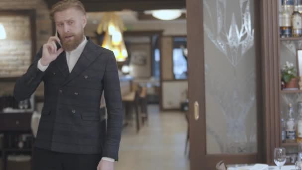 Впевнений світлий бородатий чоловік у стильній куртці, що розмовляє мобільним телефоном, стоїть у сучасному ресторані. Бізнесмен вирішує свої проблеми онлайн — стокове відео