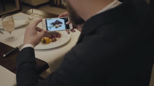 男性坐在餐厅的桌子上，拍着盘子里的大牛排的照片。从男人的背面射击. — 图库视频影像