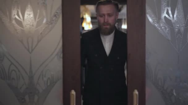 재킷을 입은 잘생긴 수염을 기른 금발 남자가 미닫이 유리문을 열고 식당에 가까이 들어와 있습니다. 뒤에 방문자를위한 테이블이있는 아름다운 편안한 방 — 비디오