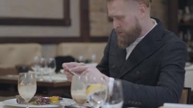 Portre kendine güvenen sarışın sakallı adam şık ceket modern bir restoranda oturan cep telefonu ile onun gıda bir fotoğraf çekerken. Yakışıklı adam kafede yalnız eğlenin