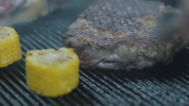 Leckere saftige Rindfleischsteaks liegen auf der Grillfläche mit Mais in Nahaufnahme. zwei rohe Fleischstücke, die in der Restaurantküche zubereitet werden. Koch wendet Fleisch mit einer Zange. der Koch bereitet schmackhaftes Essen zu — Stockvideo