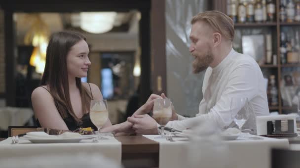 Πορτρέτο αίγλη μελαχρινή γυναίκα και ένα όμορφο μούσι ξανθό άντρα κάθεται στο τραπέζι μπροστά από το άλλο. Ο άνθρωπος που λέει καλά λόγια στην κοπέλα του, του φιλάει το χέρι. Το χαριτωμένο ζευγάρι έχει ένα — Αρχείο Βίντεο