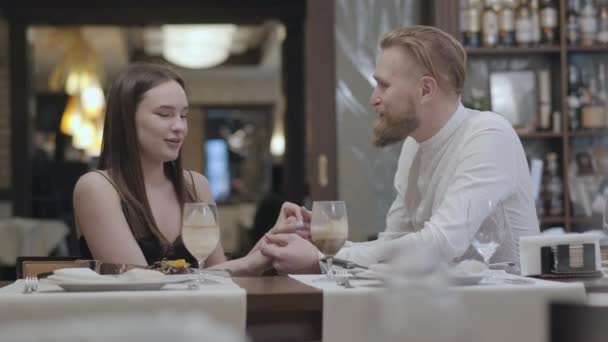 Retrato mujer morena linda y un hombre guapo barbudo rubio sentado en la mesa delante del otro. El hombre le dice buenas palabras a su novia y sostiene a la chica de la mano. La linda pareja — Vídeos de Stock