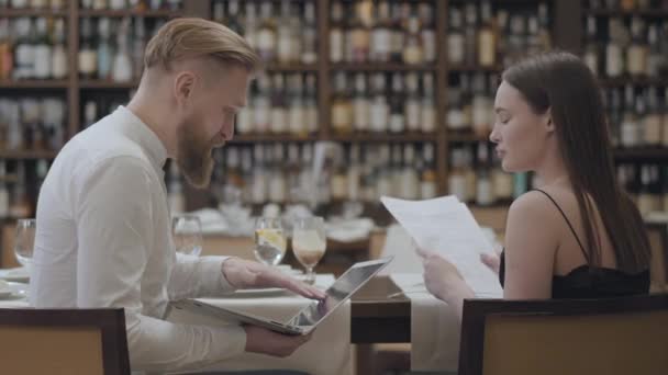 Retrato linda mujer morena y hombre barbudo rubio sentado en la mesa en el café discutiendo información en la pantalla del ordenador portátil. El hombre sosteniendo gadget. Reunión con el compañero de trabajo — Vídeo de stock