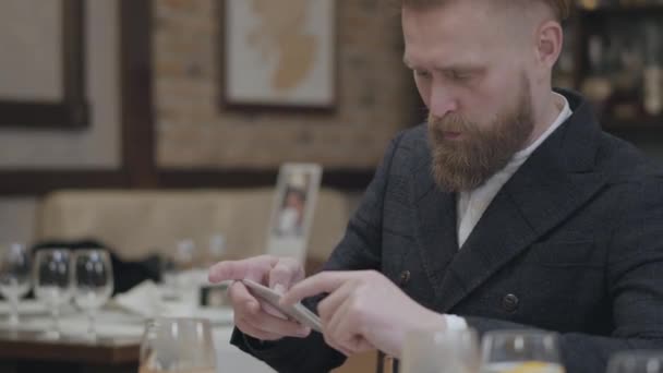 Kendine güvenen sarışın sakallı adam şık ceket modern bir restoranda oturan cep telefonu ile onun gıda bir fotoğraf çekerken. Yakışıklı adam kafede yalnız eğlenin — Stok video