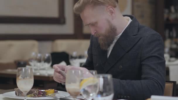 Retrato barbudo empresário de sucesso em casaco elegante tirando uma foto de sua comida com seu celular sentado em um restaurante moderno. Bonito homem se divertir no café sozinho — Vídeo de Stock