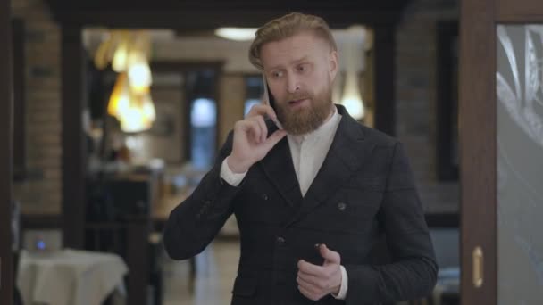 Portret van een succesvolle zaken man met een rode baard die op zijn cel praat in een duur restaurant of café. — Stockvideo