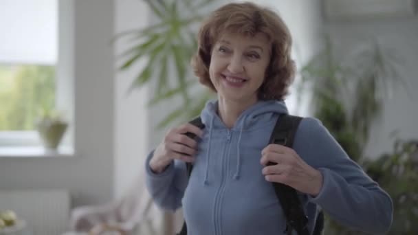 Портрет зрілої щасливої жінки в синьому халаті, що фіксує чорний зручний рюкзак на спині — стокове відео