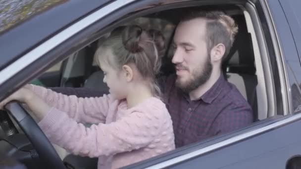 Porträtt liten flicka sitter på pappor knä i bilen närbild. Barnet lär sig att köra bilen. Leende man spenderar tid med sin dotter. Familj fritid, folk skrattar. Kamera rörliga — Stockvideo