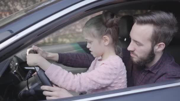 Liten flicka sitter på pappor knä i bilen närbild. Barnet lär sig att köra bilen. Leende man spenderar tid med sin dotter. Familj fritid, folk skrattar. Kamera flyttar vänster — Stockvideo
