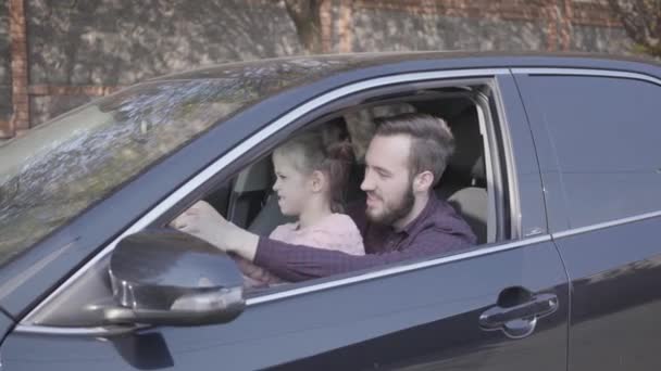 Liten flicka sitter på pappor varv i bil närbild. Barnet lär sig att köra bilen. Mannen och ungen visar stora fingrar. Leende man spenderar tid med dotter. Familje fritid, resekoncept — Stockvideo