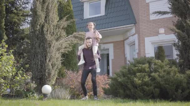 Mladý pohledný otec stojí před velkým domem s dcerou na ramenou. Dítě držící malého čivavského psa. Muž, který se krčí a dělá sport. Rodinný pronájem — Stock video