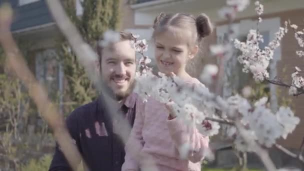 Portret jonge bebaarde vader houdt zijn kleine lachende dochter op zijn armen, zodat ze de boom bloesem close-up kon ruiken. Familie vrije tijd buitenshuis, lente — Stockvideo