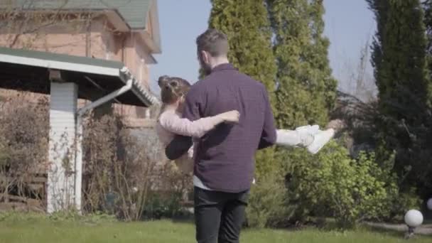 Joven padre barbudo sosteniendo a su pequeña hija sonriente en sus brazos girando, ambos riendo. Ocio familiar al aire libre, primavera, convivencia. Cámara acercándose — Vídeos de Stock