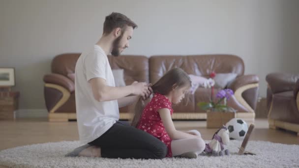 Giovane padre barbuto spazzolare i capelli della sua bambina mentre il bambino pettinare la coda del suo cavallo giocattolo seduto sul pavimento su un tappeto morbido. Tempo libero in famiglia. Paternità, cura, amore. Vista laterale — Video Stock