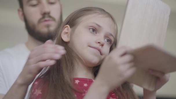 Jeune père barbu mettant la couronne sur la tête de sa petite fille, faisant de sa princesse tandis que l'enfant se regarde dans le miroir de près. Loisirs en famille. Paternité, compassion, amour. Vue du bas — Video