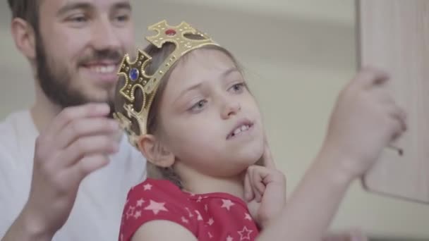 Ritratto attraente giovane padre barbuto mettere la corona sulla testa della sua bambina, facendo la sua principessa mentre il bambino si guarda allo specchio da vicino. Tempo libero in famiglia. Paternità, cura, amore — Video Stock