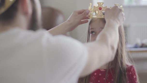 Молодой отец надевает корону на голову своей маленькой дочери, делая ее принцессу крупным планом. Девушка счастлива, она поднимается и хлопает в ладоши. Семейный отдых. Отцовство, забота, любовь — стоковое видео