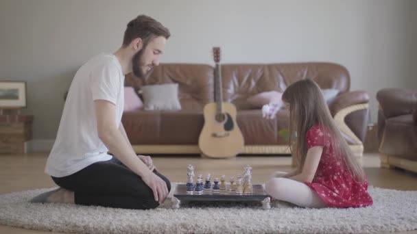 Genç sakallı baba ve uzun saçlı küçük kız deri koltuğun önünde yerde oturmuş satranç oynuyorlar. Aile eğlencesi. Babalık, çocukluk, aşk. Yan görünüm — Stok video