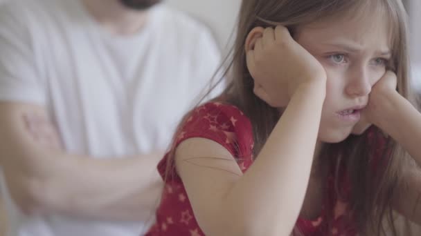 Retrato de uma menina triste sentada em primeiro plano segurando a cabeça de perto. O pai com braços cruzados no fundo. A criança ofendida pelo pai. Dificuldades em crescer crianças — Vídeo de Stock