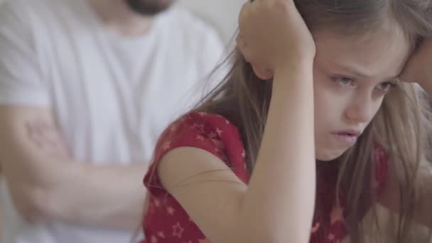 The sad little girl sitting on the foreground holding her head in hands close up. The father with crossed arms on the background. The child offended by her dad. Difficulties in growing up kids — Stock Video