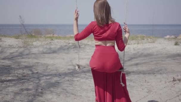 Retrato delgado mujer elegante en vestido rojo sentado en el columpio y mirando una increíble vista espectacular del lago azul en frente de ella . — Vídeos de Stock