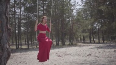 Ormanda salıncak üzerinde oturan ve inanılmaz bir muhteşem görünümü görünümlü kırmızı elbiseli zarif güzel genç kadının portresini kapatın. Yavaş çekim.