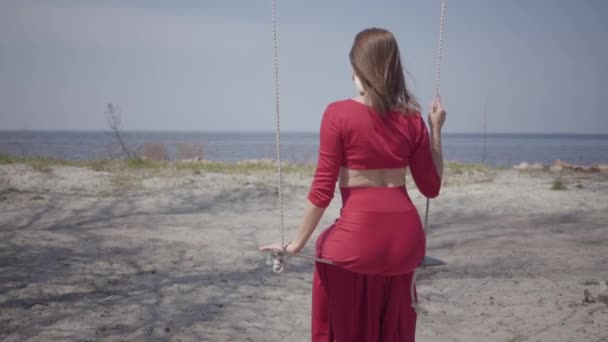 Portrait slim elegant woman in red dress sitting on the swing and looking an amazing spectacular view of blue lake in front of her. Slow motion. — Stock Video