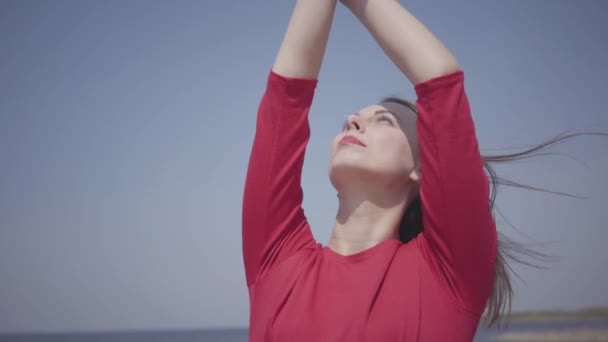 A mulher graseful do retrato na dança longa vermelha do vestido levanta as mãos na pedra grande perto da vista espetacular da água do mar azul e do céu surpreendente. Dançarina contemporânea a praticar ao ar livre. Movimento lento . — Vídeo de Stock